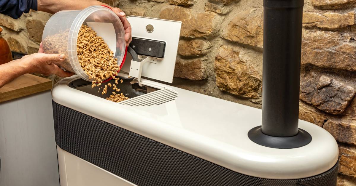 A person in a blue shirt pouring a bucket of wood pellets into a stove. The wall behind the stove is made of rocks.