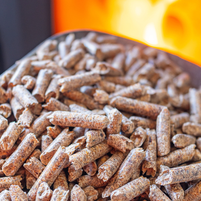 A silver scoop full of small wood pellets held in front of a lit black and red pellet stove fireplace.