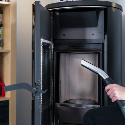 A hand holding a black and silver vacuum cleaner up to an open black pellet stove fireplace, preparing to clean it.