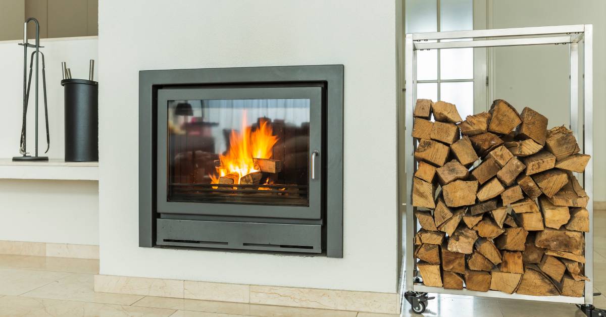 A fireplace with a glass door next to a rolling cart carrying a tall stack of chopped wood logs in a white room.