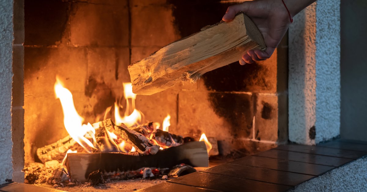 A hand adds a wood log to a small fire burning in a home fireplace. The flames of the fire are yellow.