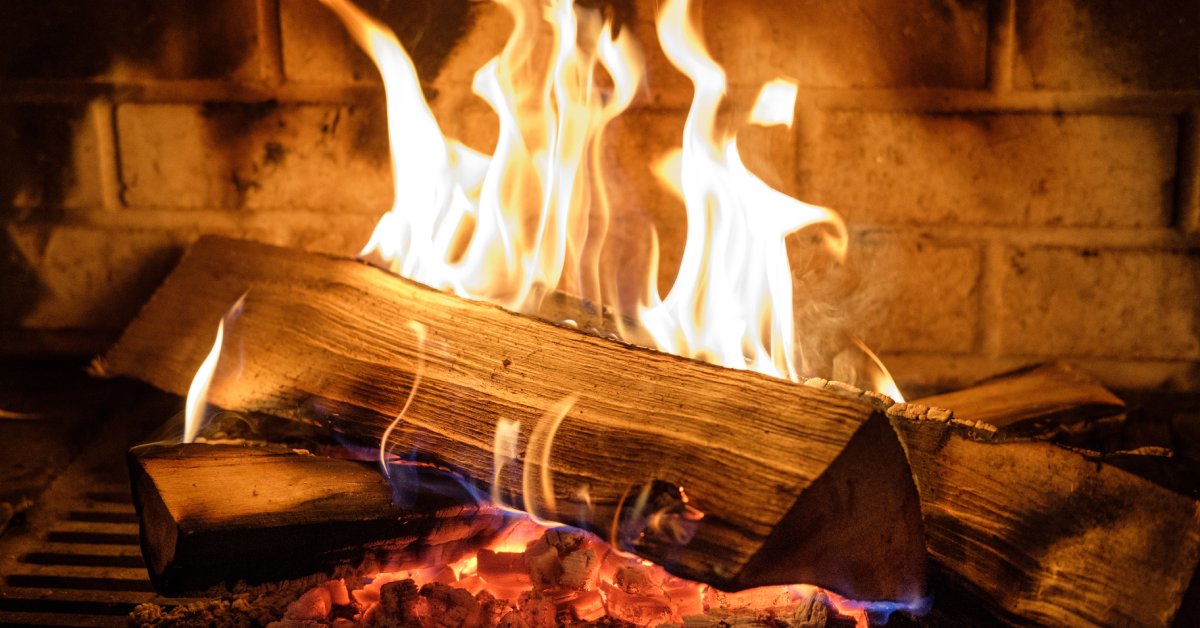 Several wood logs burn in a fire in a fireplace with a brick background. The flames are a pale yellow and blue.