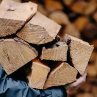 A man in a dark blue raincoat carries a stack of seven pieces of firewood past a wall of wood in the background.