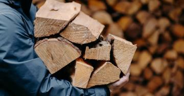 A man in a dark blue raincoat carries a stack of seven pieces of firewood past a wall of wood in the background.